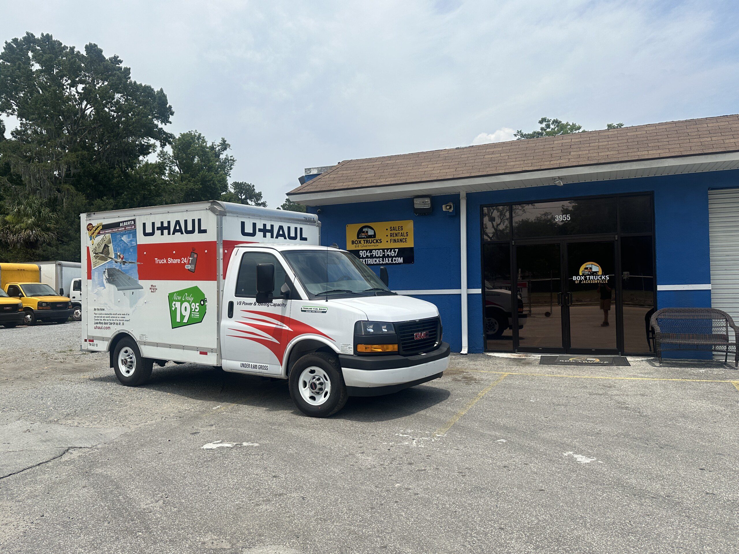 A U-Haul truck is parked in front of a building with a sign that reads "Postal Depot" and a phone number. The truck has U-Haul branding and an advertisement for $19.95 rentals. The building has a glass door and windows, with a yellow and blue sign above it.